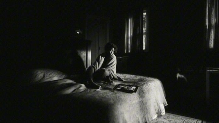 Black and white still from Un/Dress: a woman wrapped in a blanket sits alone on a bed in a dark room lit by a window.