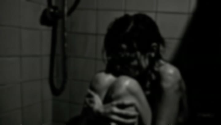 Black and white still from Un/Dress: a person crouches in a tiled shower, arms wrapped around their knees, water on their skin.